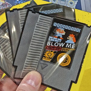May include: Three gray and black iron-on patches featuring a retro video game cartridge design. The patches are labeled "Blow Me" and "Nintendo Entertainment System". The patches are 2.9 inches wide and 3.1 inches high.