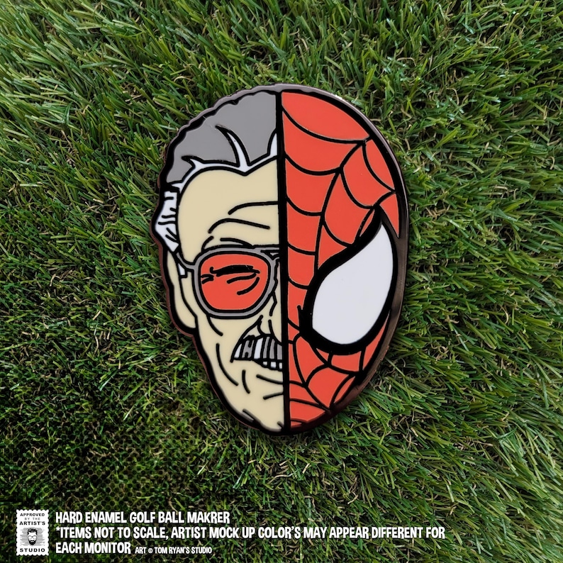 K&ouml;nnte beinhalten: Ein Golfballmarker aus Hartemaille mit geteiltem Design. Eine H&auml;lfte zeigt Stan Lees Gesicht, die andere H&auml;lfte die Maske von Spider-Man. Der Marker liegt auf einem gr&uuml;nen, grasbewachsenen Hintergrund.