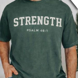 Puede incluir: Camiseta verde oscuro con la palabra "STRENGTH" en letras blancas en negrita y "PSALM 46:1" debajo. La camiseta la lleva una persona con vaqueros azules. El logotipo de la marca "feathers" está en la esquina superior derecha.