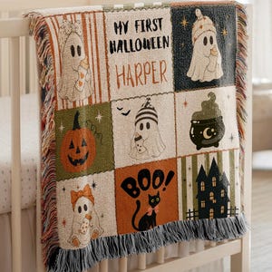 May include: A woven throw blanket featuring a grid of Halloween-themed illustrations including ghosts, a pumpkin, a black cat, a cauldron, and a haunted house. The text reads MY FIRST HALLOWEEN HARPER and BOO. The blanket has a fringed edge.