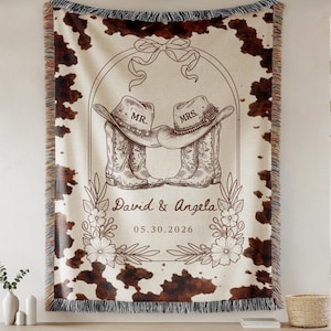 May include: A woven throw blanket featuring a brown and white cowhide print pattern. The centre displays a line art illustration of two cowboy boots with hats labelled MR. and MRS., the names David & Angela, and the date 05.30.2026 with floral accents.