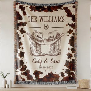 May include: A woven throw blanket featuring a brown and white cowhide print border. The center displays the text THE WILLIAMS, a horseshoe icon, two cowboy hats labeled MR. and MRS., the names Cody & Sara, and the date 10.10.2026.