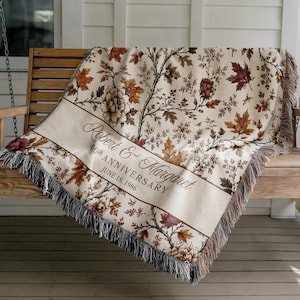 May include: A woven throw blanket featuring a cream background with a pattern of brown and orange autumn leaves and flowers. A central diagonal band displays the text Robert & Margaret, 40th ANNIVERSARY, JUNE 18, 1986 in dark brown script.