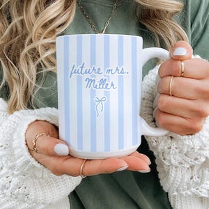 May include: A white ceramic mug with vertical light blue stripes. The mug has the text "future mrs. Miller" in light blue script, with a small bow illustration. The mug is held by a person wearing a white sweater.