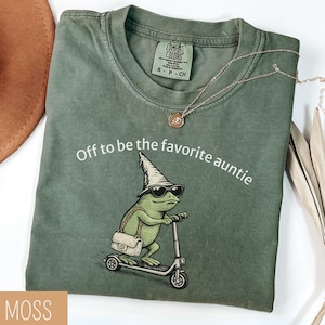 May include: A moss green t-shirt with the phrase "Off to be the favorite auntie" and a graphic of a frog riding a scooter, wearing sunglasses and a hat. A gold necklace and a brown hat are also visible.