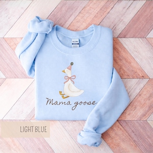 May include: Light blue sweatshirt with a watercolor illustration of a goose wearing a party hat and a pink bow. The words "Mama goose" are written in a cursive font below the goose. The bottom left corner of the image has the words "LIGHT BLUE".