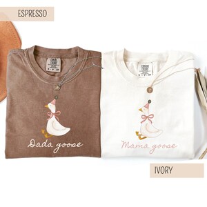 May include: Two Comfort Colors t-shirts, one espresso and one ivory, featuring a goose graphic wearing a party hat and bow. The espresso shirt says "Dada goose" and the ivory shirt says "Mama goose". Both shirts have a silver necklace.