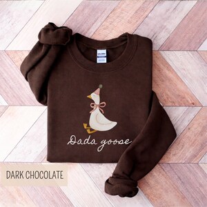 May include: Dark chocolate-colored sweatshirt featuring a whimsical illustration of a goose wearing a party hat and a pink bow. The text "Dada goose" is written in cursive below the goose. The words "Dark Chocolate" are printed at the bottom.