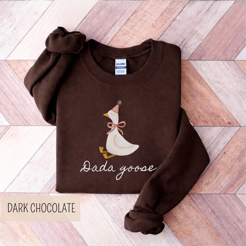 May include: Dark chocolate-colored sweatshirt featuring a whimsical illustration of a goose wearing a party hat and a pink bow. The text "Dada goose" is written in cursive below the goose. The words "Dark Chocolate" are printed at the bottom.