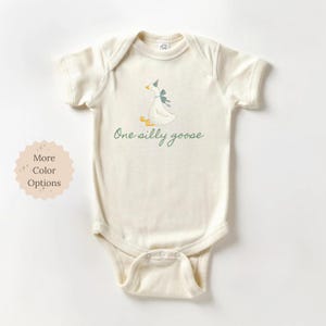 May include: Cream-colored baby onesie with a whimsical illustration of a goose wearing a party hat and bow. The text "One silly goose" is written in a script font. The onesie has short sleeves and snap closures.