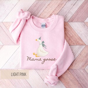 May include: Light pink sweatshirt with a graphic of a goose wearing a party hat and a bow. The text "Mama goose" is written in a script font below the goose. The words "LIGHT PINK" are in a rectangular box at the bottom.