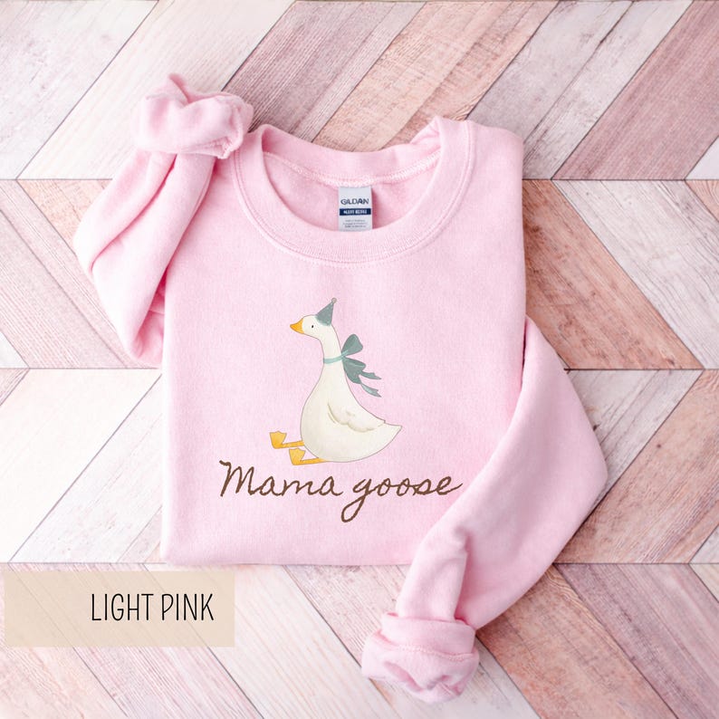 May include: Light pink sweatshirt with a graphic of a goose wearing a party hat and a bow. The text "Mama goose" is written in a script font below the goose. The words "LIGHT PINK" are in a rectangular box at the bottom.