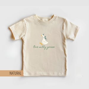May include: A natural-colored t-shirt featuring a whimsical illustration of a goose wearing a party hat and bow. The text "One silly goose" is written below the goose in a script font. The shirt is made of a soft material.