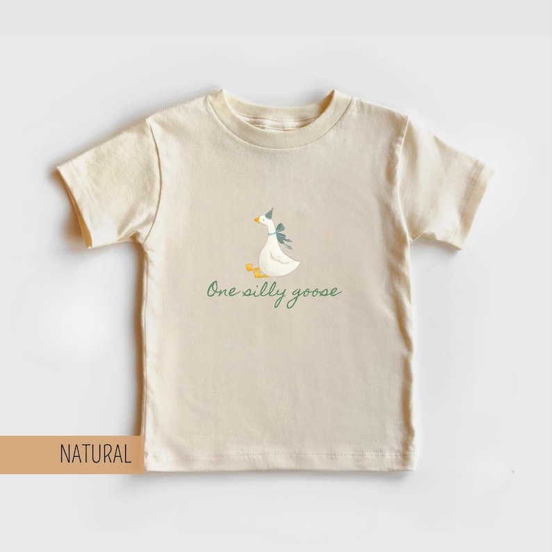 May include: A natural-colored t-shirt featuring a whimsical illustration of a goose wearing a party hat and bow. The text "One silly goose" is written below the goose in a script font. The shirt is made of a soft material.