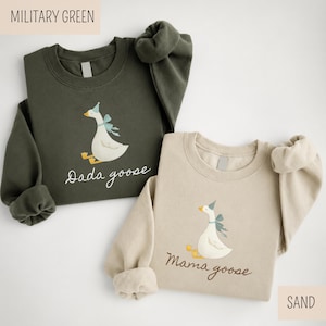 May include: Two crewneck sweatshirts, one military green and one sand-colored. Each features a whimsical goose illustration with a party hat and bow. The military green sweatshirt says "Dada goose," and the sand-colored one says "Mama goose."