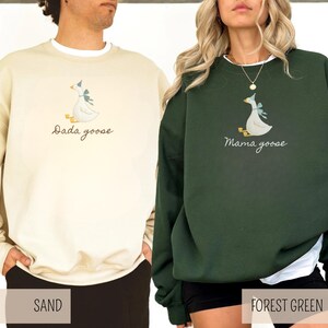May include: Two sweatshirts, one sand-colored and one forest green, each featuring a goose graphic with a blue bow. The sand-colored sweatshirt has the text "Dada goose," while the forest green one says "Mama goose."