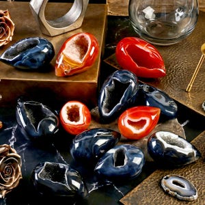 May include: A collection of polished agate geodes in shades of blue, red, and orange. Some geodes are cut open, revealing crystal interiors. The arrangement includes a silver ring and a clear glass vase, set on a textured surface.