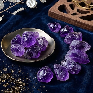 May include: A collection of raw amethyst crystals in varying sizes and shapes. The deep purple stones are displayed on a small, antique-looking metal dish and scattered on a dark blue velvet surface. A wooden decorative piece is in the background.