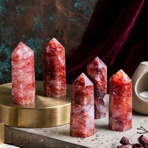 Puede incluir: Cinco torres de cristal de cuarzo rojo con vetas naturales. Los cristales se exhiben sobre un soporte dorado y una superficie de hormigón gris. Los cristales tienen una parte superior facetada y pulida y un cuerpo cilíndrico.