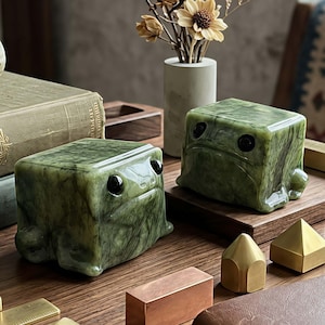 May include: Two cube-shaped green stone figurines with smooth, marbled textures and dark, glossy circular eyes. These blocky, frog-like sculptures feature rounded corners and a polished finish, positioned alongside various geometric metal and wooden objects.