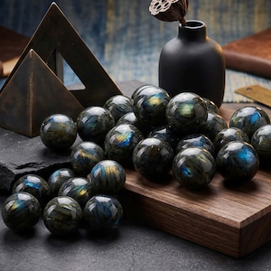 以下が含まれることがあります： 青、緑、金色の虹色の輝きを放つ、研磨されたラブラドライトの球体のコレクション。 球体は木製の表面に配置され、背景には暗い花瓶と幾何学的な金属彫刻があります。