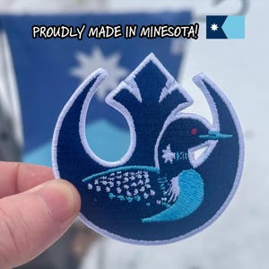 May include: A navy blue embroidered patch featuring a loon design with a red eye and turquoise accents. The patch is in a circular shape with a white border. Text above the patch reads "PROUDLY MADE IN MINNESOTA!"