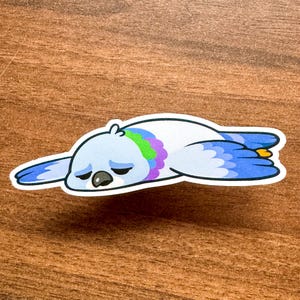 May include: A sticker featuring a cartoon pigeon lying down, with a sad expression. The pigeon is light blue with a white belly, and has a colorful green and purple neck. The wings and tail are shades of blue.