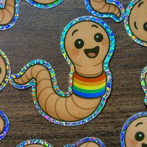 May include: A collection of stickers featuring a smiling cartoon worm with a rainbow-colored collar. The worm is light brown with a glittery, iridescent border. The stickers are on a dark brown surface.