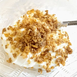 May include: A clear glass bowl filled with white yogurt topped with a generous amount of crushed, golden brown cereal.
