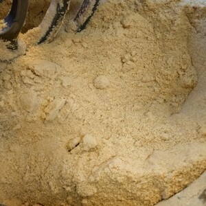 May include: A close-up of a beige powder in a mixing bowl with a metal mixer attachment. The powder is evenly distributed throughout the bowl.