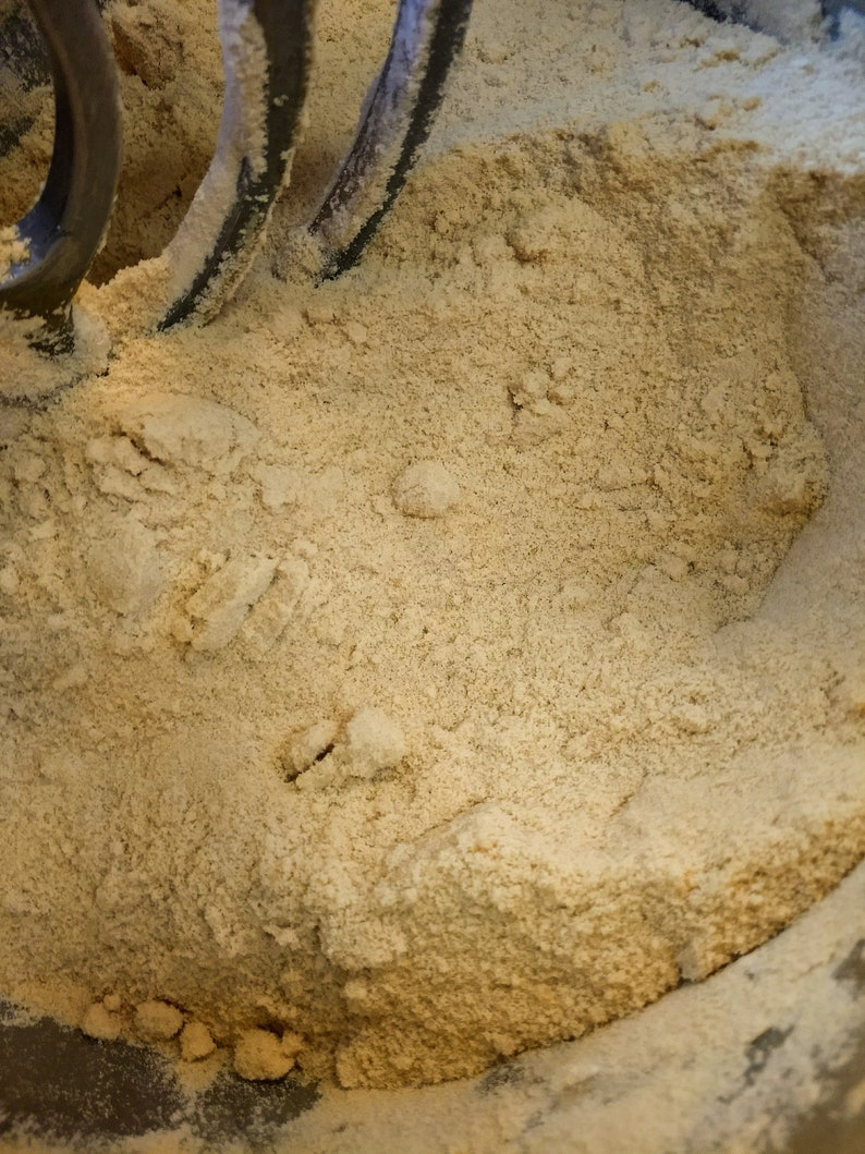 May include: A close-up of a beige powder in a mixing bowl with a metal mixer attachment. The powder is evenly distributed throughout the bowl.