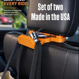 May include: An orange plastic car headrest hook featuring a molded American flag design. The hook is attached to a black leather car headrest and holds black fabric straps. Text on the image reads: MORE SPACE. LESS MESS. EVERY RIDE. Set of two Made in the USA.