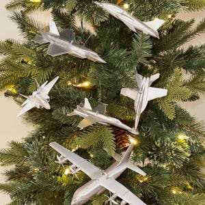 May include: A collection of silver-colored airplane ornaments hanging on a Christmas tree. The ornaments depict various aircraft, including fighter jets and a propeller plane. The tree is decorated with lights and pine needles.