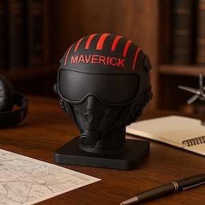 Peut inclure: Un casque décoratif noir avec des détails rouges et le mot "MAVERICK" en haut. Le casque, ressemblant à un casque de pilote, est posé sur une base noire. L'image est sur un bureau en bois avec une carte, un carnet, un stylo et un casque.