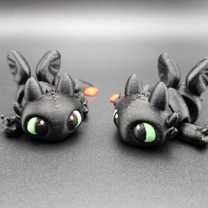 Könnte beinhalten: Zwei schwarze Drachenfiguren mit grünen Augen und orangefarbenen Akzenten. Die Drachen liegen, mit ausgebreiteten Flügeln und kleinen Füßen. Die Figuren haben eine strukturierte Oberfläche und sind vor einem dunklen Hintergrund platziert.