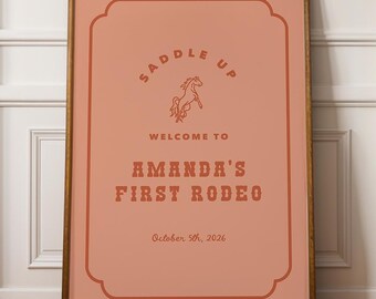 May include: A framed poster with a peach background and brown text. The text reads "SADDLE UP" above a horse illustration, and "WELCOME TO AMANDA'S FIRST RODEO" below. The date "October 5th, 2026" is at the bottom.