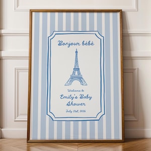 May include: A framed baby shower sign with a blue and white striped background. The sign features the Eiffel Tower and the words "Bonjour bébé" and "Welcome to Emily's Baby Shower July 21st, 2026" in blue script.