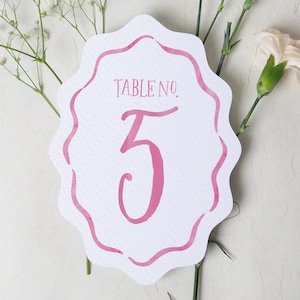 May include: A white table number card with a pink scalloped border and the number "3" in pink. The card reads "TABLE NO." above the number. The card is decorated with white flowers and greenery.