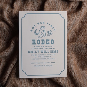May include: A baby shower invitation with a rodeo theme. The invitation is white with a blue border and text. The text includes "Not Her First Rodeo" and details for Emily Williams' baby shower. The invitation is on a brown textured surface.