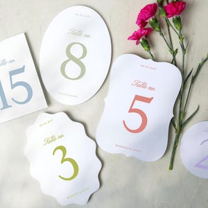 May include: A collection of white table number cards in various shapes, each displaying a different number in soft colors. The numbers are 2, 3, 5, 8, and 15. The cards are arranged with pink flowers.
