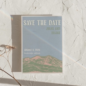 May include: A rectangular Save the Date card with a light blue background featuring a mountain range illustration in green and pink. The text reads SAVE THE DATE, JULES AND ELIJAH, AUGUST 8, 2026, and breckenridge, colorado in white and green.