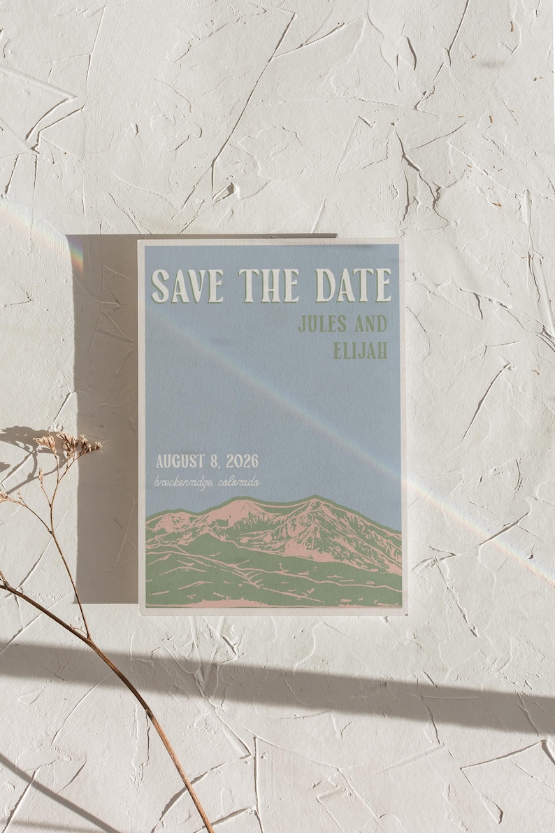 May include: A rectangular Save the Date card with a light blue background featuring a mountain range illustration in green and pink. The text reads SAVE THE DATE, JULES AND ELIJAH, AUGUST 8, 2026, and breckenridge, colorado in white and green.