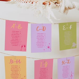 May include: A display of colorful wedding seating cards, each with a letter designation and guest names. The cards are pink, purple, and yellow, with handwritten names and decorative illustrations like champagne glasses and rings. They are clipped to a string.