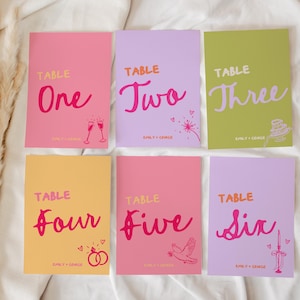 Pode incluir: Seis cartões coloridos com números de mesa com as palavras "Table One" a "Table Six" em uma fonte manuscrita. Cada cartão tem um fundo de cor diferente, incluindo rosa, roxo e amarelo. Os cartões têm ilustrações de taças de champanhe, um bolo e alianças.