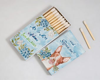 May include: A matchbox set featuring blue watercolor floral designs. One box is open, revealing white-tipped matches. Text reads Mr & Mrs Adams June 20, 2026 and and Rosie too! alongside a brown and white dog illustration. Two loose matches lie nearby.