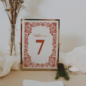 May include: A gold framed table number 7 sign with a red floral border. The sign reads "table no." above the number 7. A vase of dried flowers and a candle in a glass holder are in the background.
