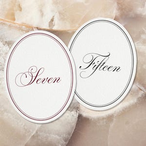 May include: Two oval-shaped white paper cards with decorative borders. The left card features the word Seven in dark red script, while the right card displays the word Fifteen in black script. Both cards have a double-line border design.