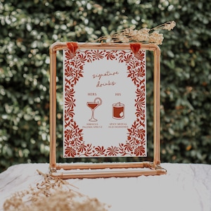 Könnte beinhalten: Ein kupferfarbener gerahmter Schild mit weißem Hintergrund und rotem Blumendesign. Auf dem Schild steht "signature drinks" und es werden zwei Cocktail-Optionen aufgeführt: Hibiscus Paloma Spritz und Spicy Mezcal Old Fashioned. Das Schild ist mit Trockenblumen verziert.
