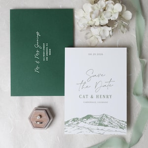 May include: A white Save the Date card with mountain illustrations, the text Save the Date, Cat & Henry, 08.29.2026, and Carbondale, Colorado. Includes a dark green envelope with Mr & Mrs Jennings 125 West Street Los Angeles, CA 90210, and two rings.