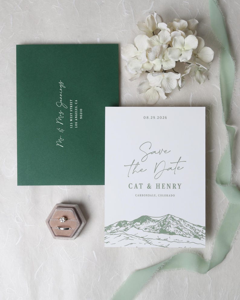 May include: A white Save the Date card with mountain illustrations, the text Save the Date, Cat & Henry, 08.29.2026, and Carbondale, Colorado. Includes a dark green envelope with Mr & Mrs Jennings 125 West Street Los Angeles, CA 90210, and two rings.